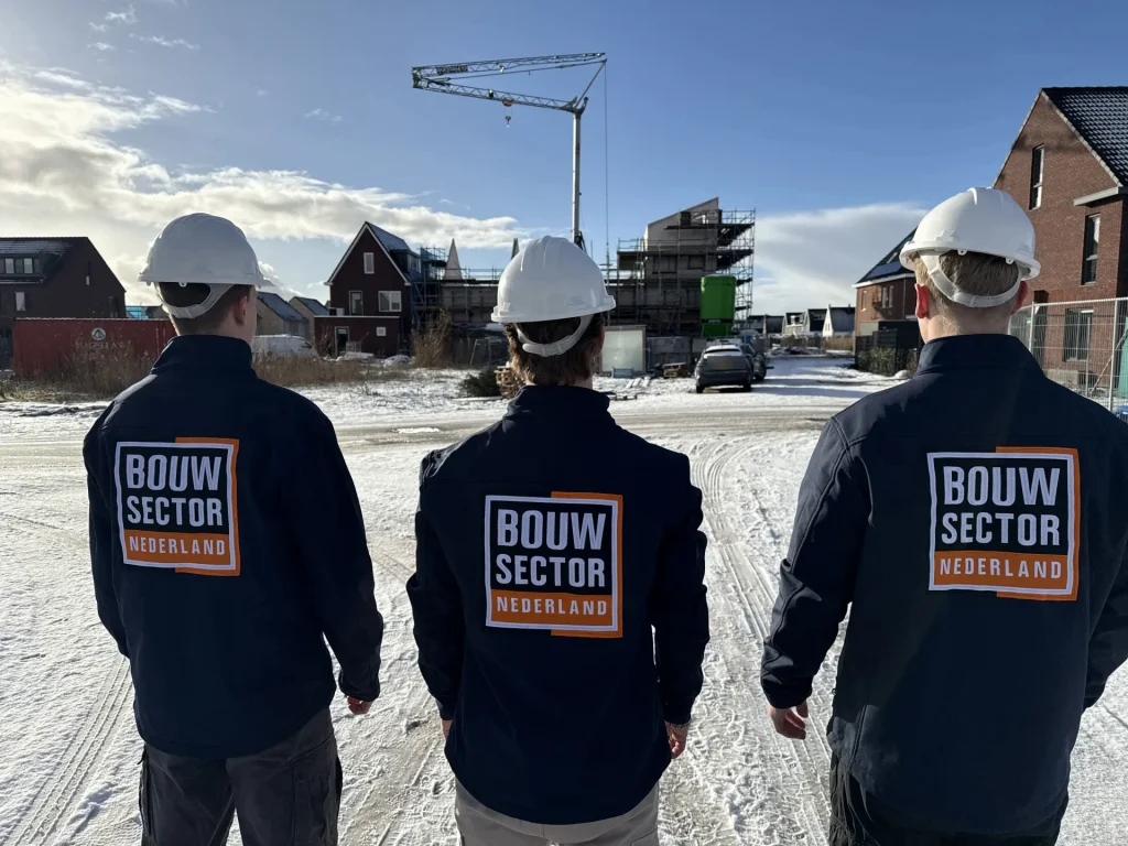 Drie vakmensen in donkerblauwe jassen met logo van Bouwsector Nederland kijken naar een nieuwbouwwijk